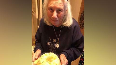 Vera Croghan, holding a plate of food and smiling to camera. She is wearing a black jumper with flowers on. 