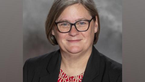 Angela Scott, a woman with glasses and short grey hair, smiles wearing a dark blazer and patterned top