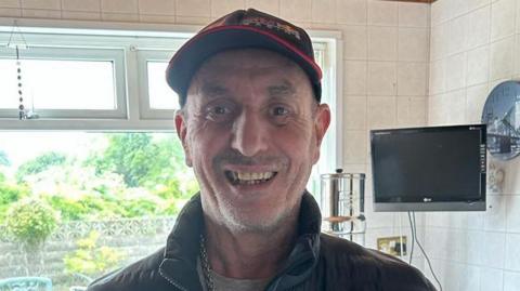 Llun o Byron Long. Mae'n gwisgo cap du a choch, crys-t llwyd a chôt ddu. Mae'n sefyll mewn cegin sy'n edrych allan ar ardd. Mae teledu a chloc ar y wal y tu ôl iddo hefyd.