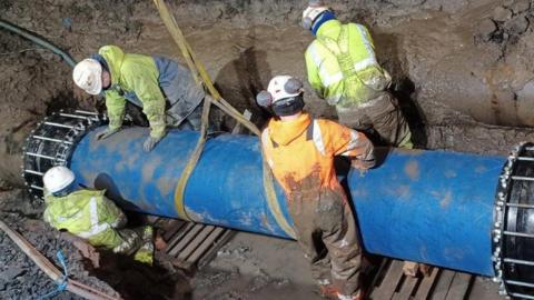 Four engineers wearing muddy fluorescent waterproofs and white helmets as they work to repair the burst water main on Pollokshaws Road in Glasgow. They are in a ditch securing a large blue pipe section, which has the yellow sling used to lower it still attached.