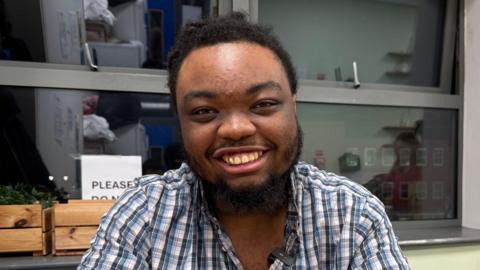 Tristan is smiling at the camera. He is black and has a beard and afro-style hair. He is wearing a blue, white and red chequered shirt. There is a window behind him and we can see it is night time.
