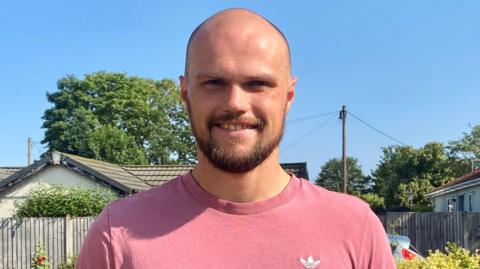 James Russell wear pink T-Shirt and is smiling. He has a beard and is looking at the camera. 