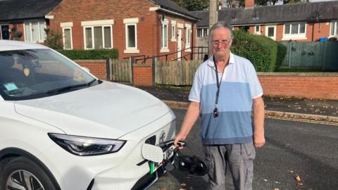 David Kelsall stands next to his electric vehicle holding a charger into the charging port at the front of the white car. He is frowning. 