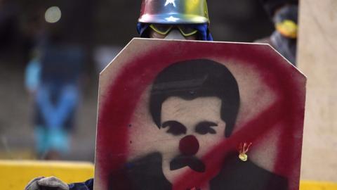 An opposition activist wearing protective helmet and gloves and a gas mask, takes cover behind a makeshift shield decorated with a scornful image of Venezuela's President Nicolas Maduro during a protest in Caracas
