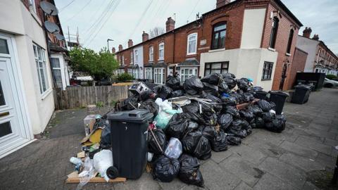 A huge pile of bin bags on a residential street. There are dozens of refuse sacks with terraced homes in the background. 