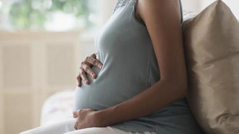 A pregnant woman sitting down cradling her bump. She is wearing a grey, sleeveless top and you cannot see her head.