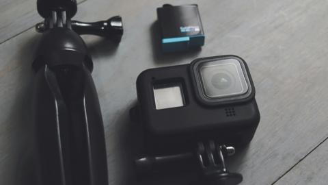 A cube shaped black camera is positioned on a grey wooden surface with a tripod and another piece of equipment that is also black and square.