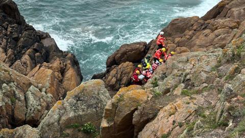 Nine emergency service workers wearing high-vis uniform and helmets are grouped together in a gap between some rocks. The cliffside is steep and rocky. The sea is a short distance from the group and it is choppy.