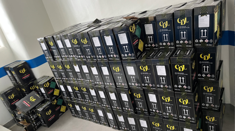 Four rows of black boxes, which are stacked two boxes deep, are lined up against a white will with a blue stripe running along the middle. The boxes have white markings and a yellow logo which reads: "CDL". There are more boxes on in the far left corner of the room.