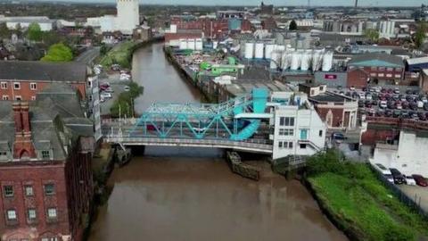 Aerial view of North Bridge. The main blue steel structure spans a muddy river with a large white building on the righthand side. Industrial buildings are visible in the background.