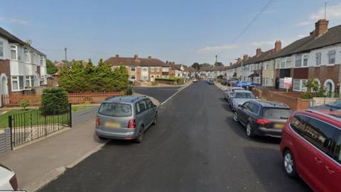 Broad Avenue with the junction to Gloucester Road on the left. It is lined with 1930s terraced houses, with small front gardens and bay windows. There are about seven cars parked along the road and pavement on either side. The road looks recently tarmacked as it is a dark grey. 