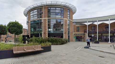 A modern building in a circular shape in brick and glass with trees and green spaces outside and benches and paved walkways