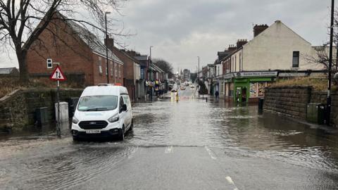 A white Northumbrian Water van parked on the side of the flooded street. A section of the road, and the surrounding pavement, is covered in water. A passer-by is standing outside Sultan Halal Superstore and looking at the flood.