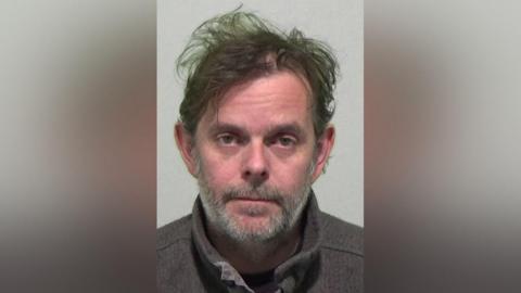 Mugshot of William Henderson who messily ruffled dark hair and a grey beard.
