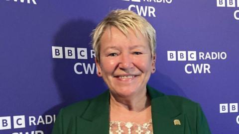 A woman with short blonde hair looks into the camera with a smile which shows her teeth. She wears a dark green jacket with a gold lapel bin in a square design. Behind her is a purple board with "BBC Radio CWR" written on it in white.