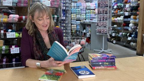 Penny, a woman with dark blonde hair, sits at a desk with a pile of books next to her, holding an open book which she is reading while smiling. In the background of the shop she is sitting in you can see shelves of yarn.