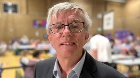 Steve Jarvis a man with black rimmed glasses is looking directly at the camera. He is wearing a black suit jacket and white shirt and is standing in a hall.