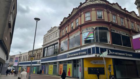 The former Co-op department store building on Carr Street, Ipswich. Shoppers are walking along the pedestrianised street past the empty store which stands on the corner. The sky is grey and overcast.