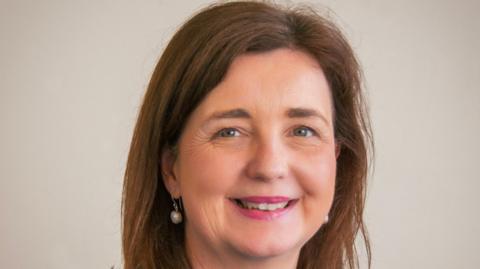 Tanya Miles, a headshot of a woman with straight brown hair, pearl earrings and pink lipstick is smiling at the camera against a plain beige background
