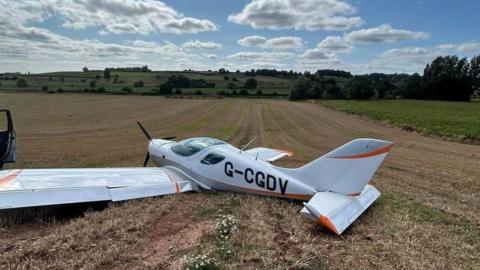 A small plane lies flat and broken in a field. It is white with orange detailing on the wing tips. It has a registration number in black lettering on the side.