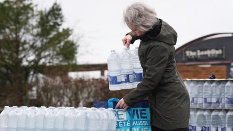 A woman places a case of bottled water into a shopping bag. The bag is held up on a large pile of water cases.