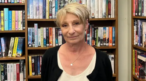 A smiling Jennie Storey looks directly at the camera as she stands in front of a bookshelf full of titles. She is wearing a black cardigan and a white shirt underneath it. She is also wearing a silver necklace with a pendant. 