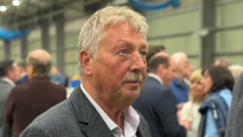 A photo of Sammy Wilson at an election count in 2024.  He has short grey hair and a grey moustache.  He is wearing a grey suit with a subtle checked pattern and an open-necked white and grey striped shirt.  Crowds of people are talking in groups in the background.