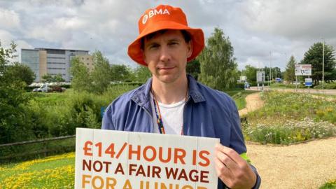 Andy Tait holding a sign saying '£14 is not a fair wage' and wearing an orange BMA hat