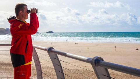 The image shows a man standing on a raised seaside viewing platform, looking out over a wide sandy beach and the sea. He is wearing a bright red jacket and red shorts with black and yellow detailing. The letters RNLI are visible on the sleeve of the jacket, along with Lifeguard text on the shorts.