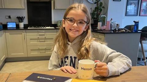 Poppy sits at her kitchen table wearing a Mensa hoodie jumper and holding a Mensa mug