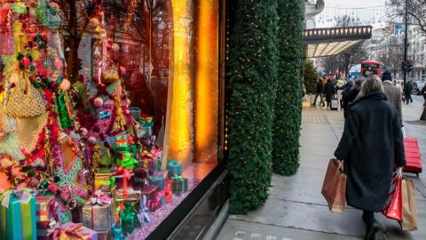 Christmas shoppers in London. A window display is packed with Christmas trees and decorative presents wrapped in shiny paper and tied with ribbons. Outside, a shopper dressed in a warm coat carries shopping bags. 