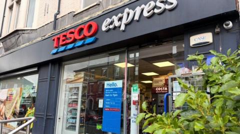 The exterior of a Tesco Express store doorway with green foliage next to it. 