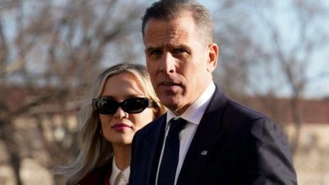 Hunter Biden in a dark suit with a dark tie with wife Melissa Cohen Biden who is wearing sunglasses in front of bare tree branches