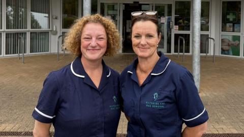 Two women in blue nurse-style uniforms stand outside a building and face the camera