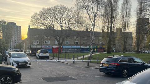 A suburban street with low rise buildings and cars parked on both sides of the road. There are trees in and green hedges and grass. A block of flats is visible at the end of the street.