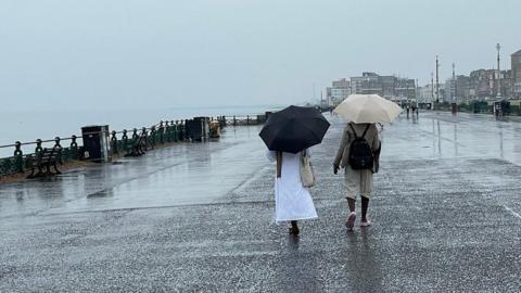 Two people with umbrellas up and their backs to the camera walk down along the Brighton seafront. The sea to the left, the sky and the pavement are all a medium shade of grey.