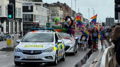 A police car leads a parade with rainbow flags seen and participants following in an open top car though the town centre