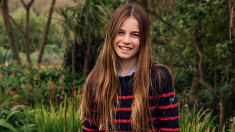Photograph shows Charlotte wearing a black and red striped jumper and blue denim jeans, standing in a flowery field while smiling at the camera.