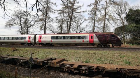 The cab and first carriage of a CrossCountry train travels on tracks in the countryside. In the background trees and fields can be seen.