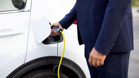 A person, whose face is out of frame, is charging a white electrical vehicle. They are wearing a navy suit.