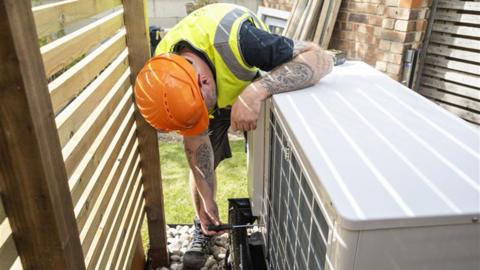 A man wearing a hard hat and high vis inspects a heat pump.