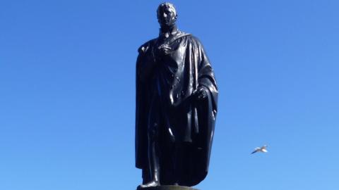 A large statue of a man standing, with a seagull flying past behind it.