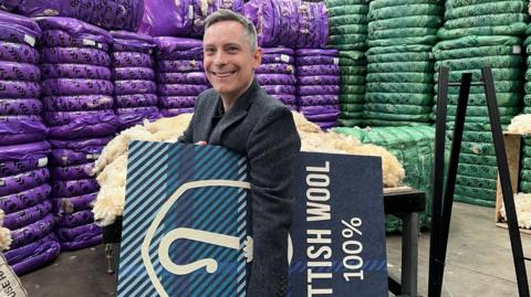 A man holding a logo board smiles at the camera, with colourful bales in the background