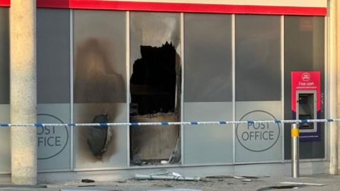 A close up of the post office on Midsummer Boulevard, Milton Keynes. It shows one of the ground-length windows has been smashed and a blackened interior. Police tape is rapped along the front of the building. 