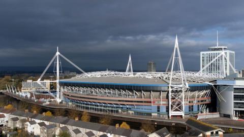 An aerial view of the Principality Stadium. Stormy grey skies are seen behind it, while in front runs the river and trees and a row of terraced houses. 