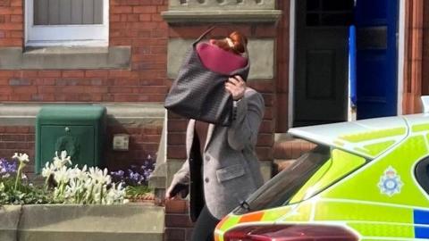 PC Alisha Staves walking on the pavement outside a red-brick building, partially covering her face with a large bag. In the foreground, the rear of a marked police car with high-visibility blue and yellow markings is parked on the street. Behind Alisha Staves are steps leading to a doorway with a blue door, a low brick wall with flowers, and a green utility box set against the building.