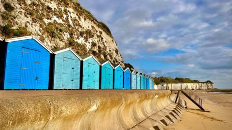 Partly sunny skies over a row of blue beach huts at the bottom of a cliff on a sandy beach