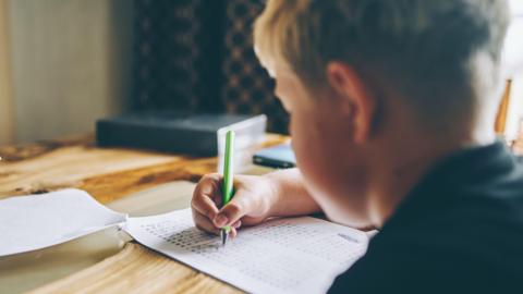 A boy doing school work at a kitchen table. He's holding a green pen. The camera is behind him. He has fair hair and is wearing a black top. 