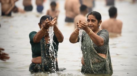 Two women wearing green sarees and gold bangles with their hair tied in a bun take a dip in the sacred waters of Sangam, the confluence of Ganges, Yamuna and mythical Saraswati rivers during the Maha Kumbh Mela festival in Prayagraj 