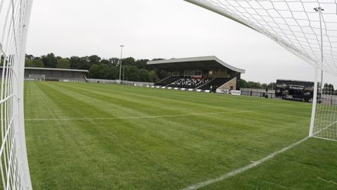 A wide angle view of Corby Town's Steel Park stadium from inside the corner of one of the goals. The green pitch is framed by a small all-seated stand with a curved roof and a terraced end behind the opposite goal.
